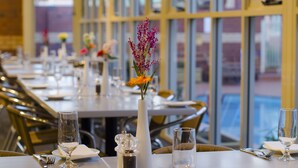 Un comedor con mesas dispuestas para el servicio, un jarrón con flores y vistas amplias por las ventanas.