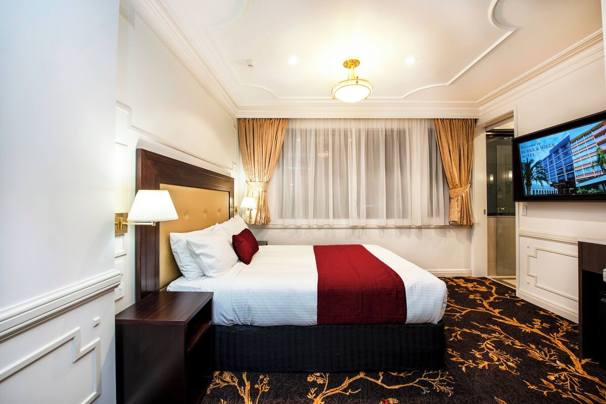 A hotel room with a neatly made bed, a television, and a large window with curtains.