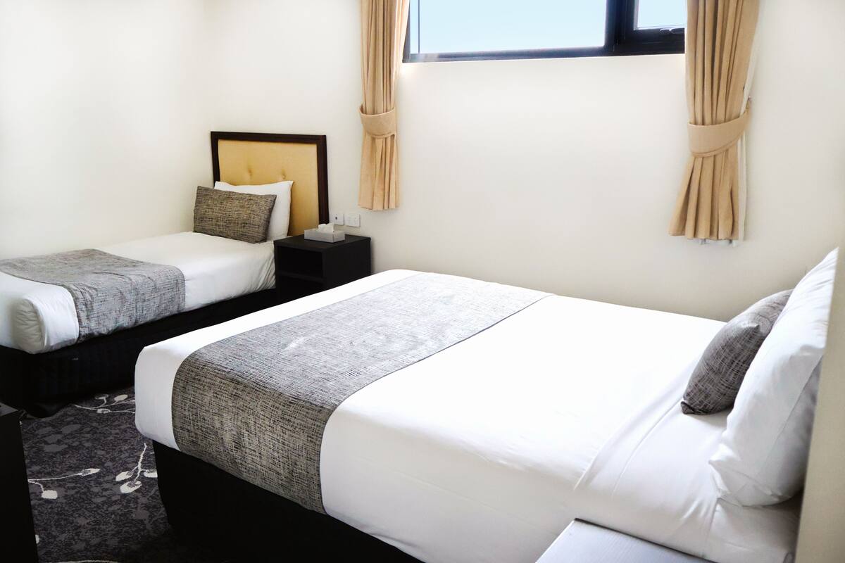 A hotel room with two beds, a nightstand, a window with curtains, and a carpeted floor.