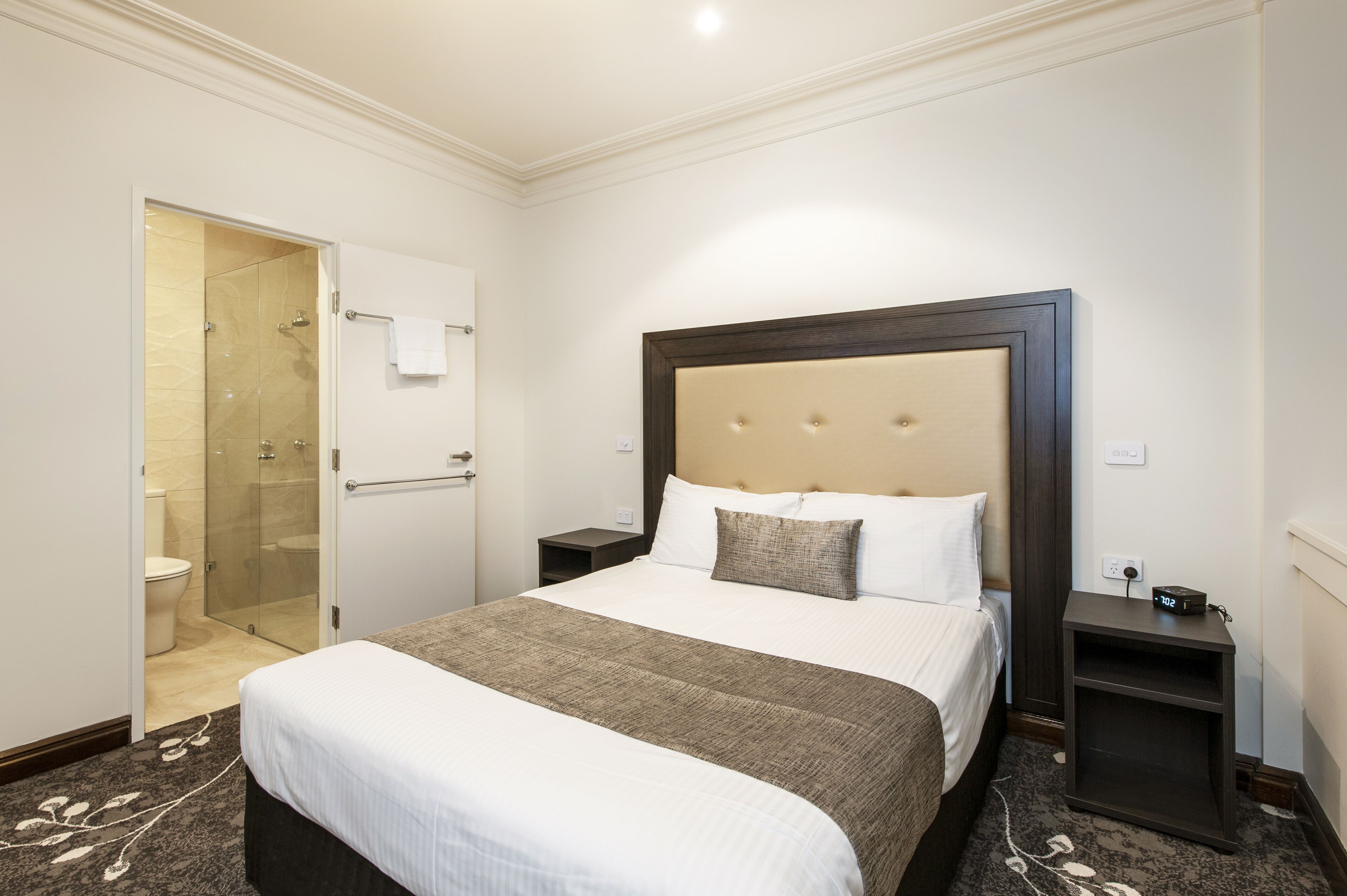 Una habitación de hotel con una cama grande, una mesita de noche, una ducha con mampara de vidrio y una puerta blanca.