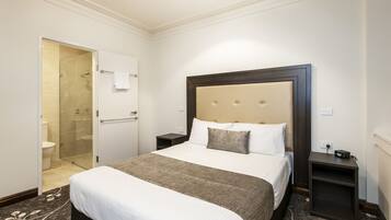 Una habitación de hotel con una cama grande, una mesita de noche, una ducha con mampara de vidrio y una puerta blanca.