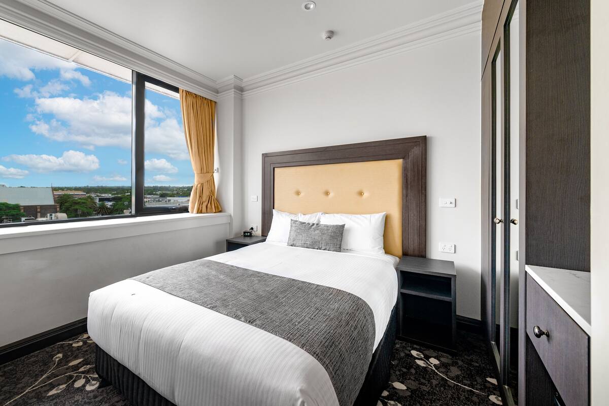 A hotel room with a large bed, a nightstand, a wardrobe, and a window with a view of the city.