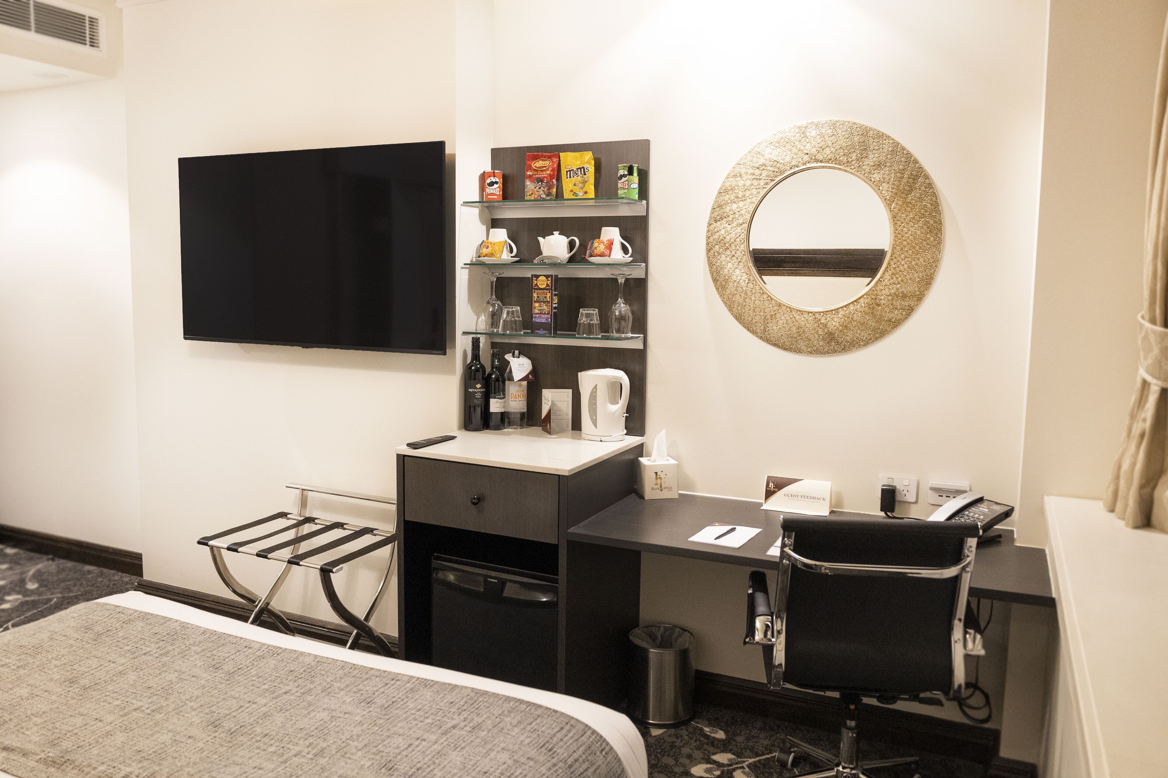 Una habitación de hotel con cama, escritorio con silla, televisión, minibar, espejo y un pequeño cubo de basura.