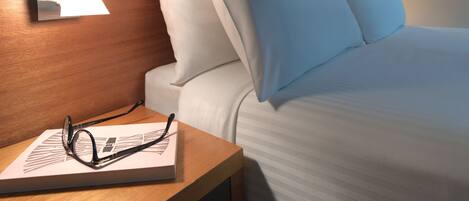 Una cama bien hecha con sábanas y almohadones blancos, una mesita de noche con una lámpara y un par de gafas sobre un libro.