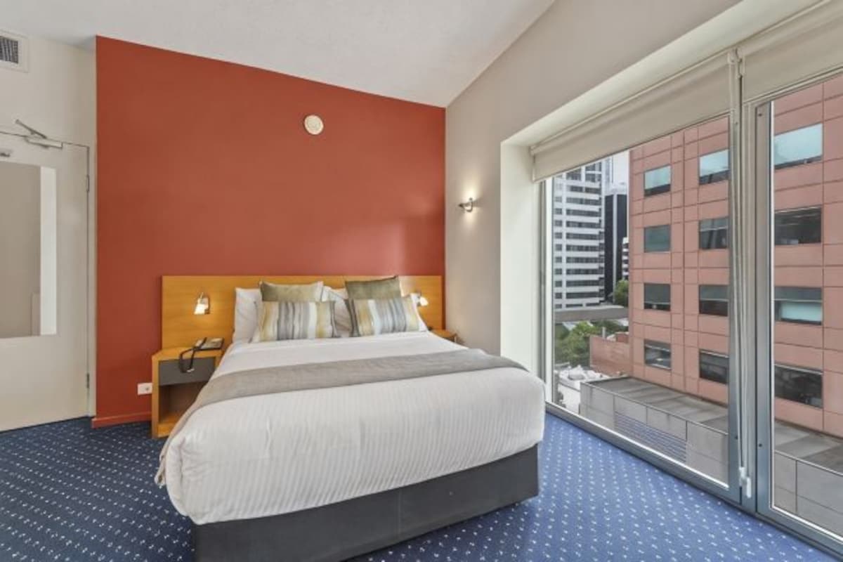 A hotel room with a large bed, a nightstand, a phone, and a window with a view of buildings.