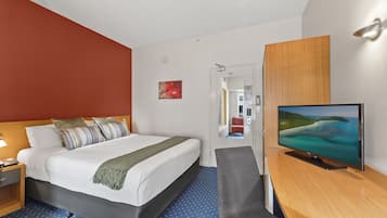 A hotel room with a bed, a desk with a flat-screen TV, a chair, a nightstand, and a red accent wall.
