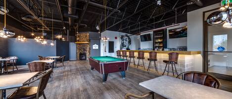 Una mesa de billar en un moderno bar o sala de estar con suelo de madera, taburetes de bar y lámparas colgantes.
