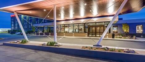 A modern hotel entrance with a covered walkway, large windows, and a visible parking area.