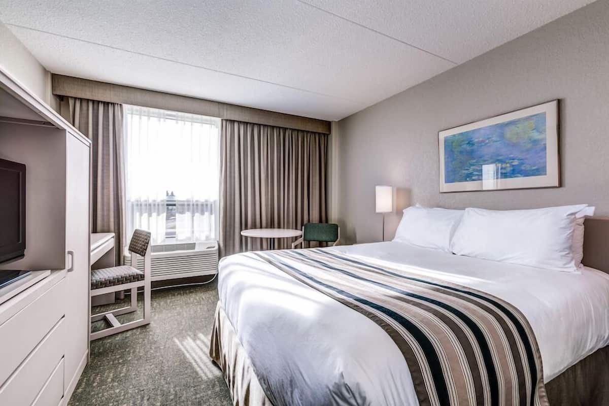 Une chambre d’hôtel avec un lit, un bureau, une chaise, une petite table, une lampe et un tableau au mur.