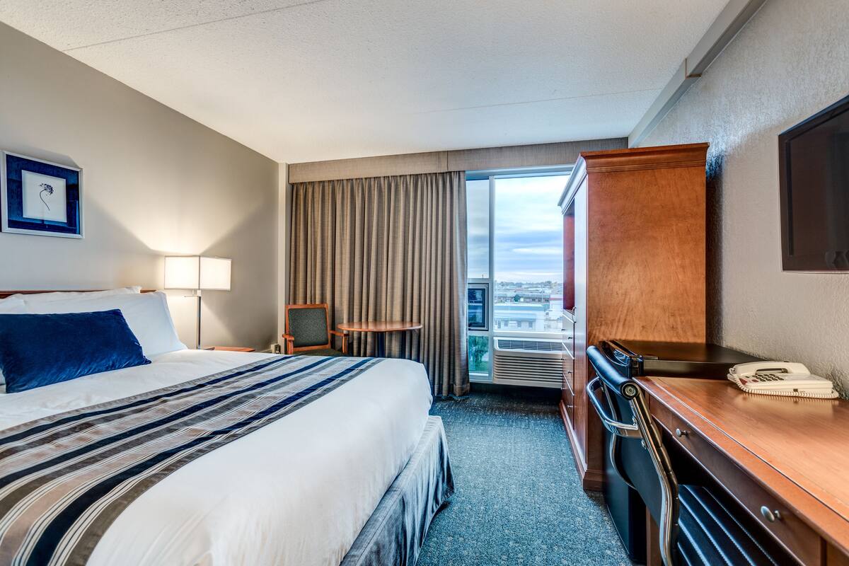 Une chambre d’hôtel avec un grand lit, un bureau, une chaise, une lampe, un téléphone et une fenêtre avec des rideaux.