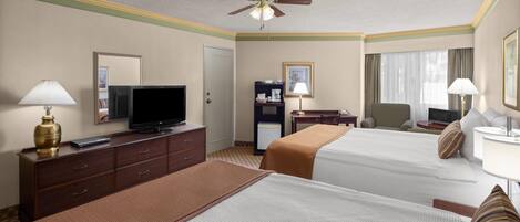 Habitación de hotel con dos camas, televisión, escritorio, silla, lámpara de escritorio, ventilador de techo y ventana con cortinas.
