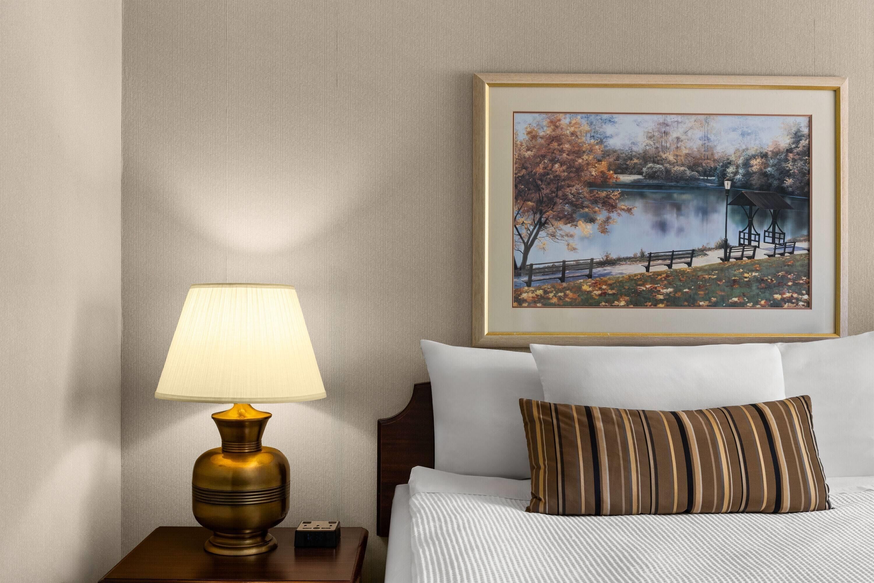 Un lit bien fait, avec un oreiller à rayures, une lampe de chevet et, au mur, un tableau représentant un paysage au bord d’un lac.