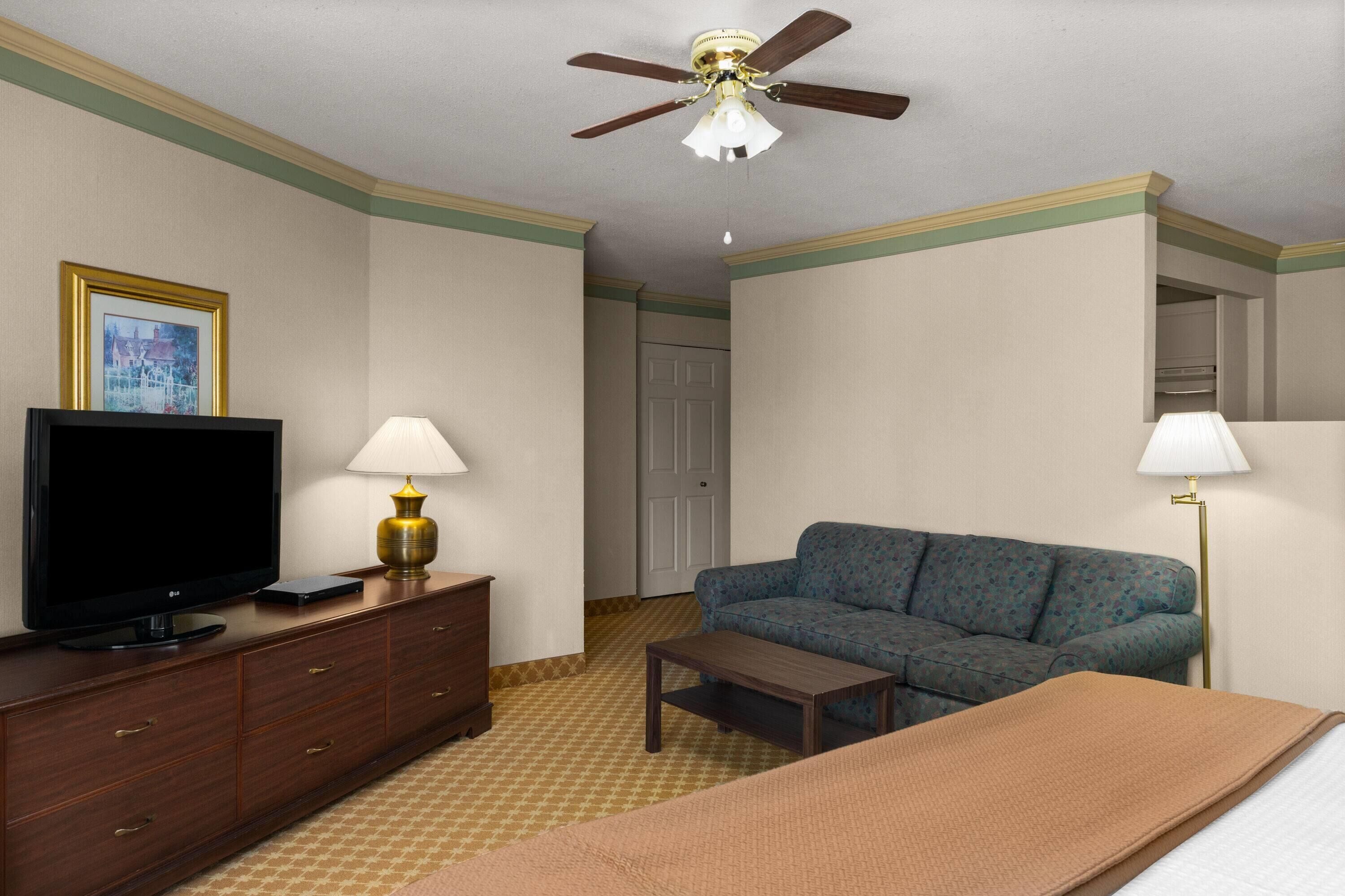 Una habitación de hotel con televisión, dos lámparas, un sofá, una cama, ventilador de techo y una puerta.