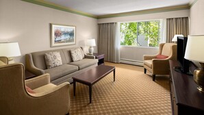 A hotel room with a sofa, armchairs, a coffee table, a TV, a lamp, a painting, and a window with curtains.