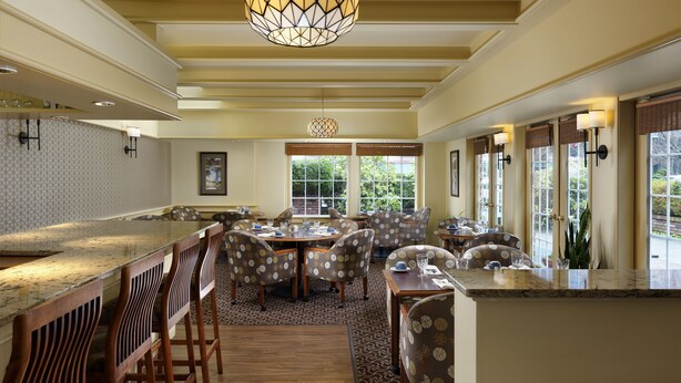 A spacious dining area with a central table surrounded by patterned chairs, a bar with a marble countertop, and a large geometric pendant light.