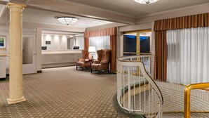 A hotel lobby with a patterned carpet, a reception desk, two armchairs, a staircase with a railing, and a large window with curtains.