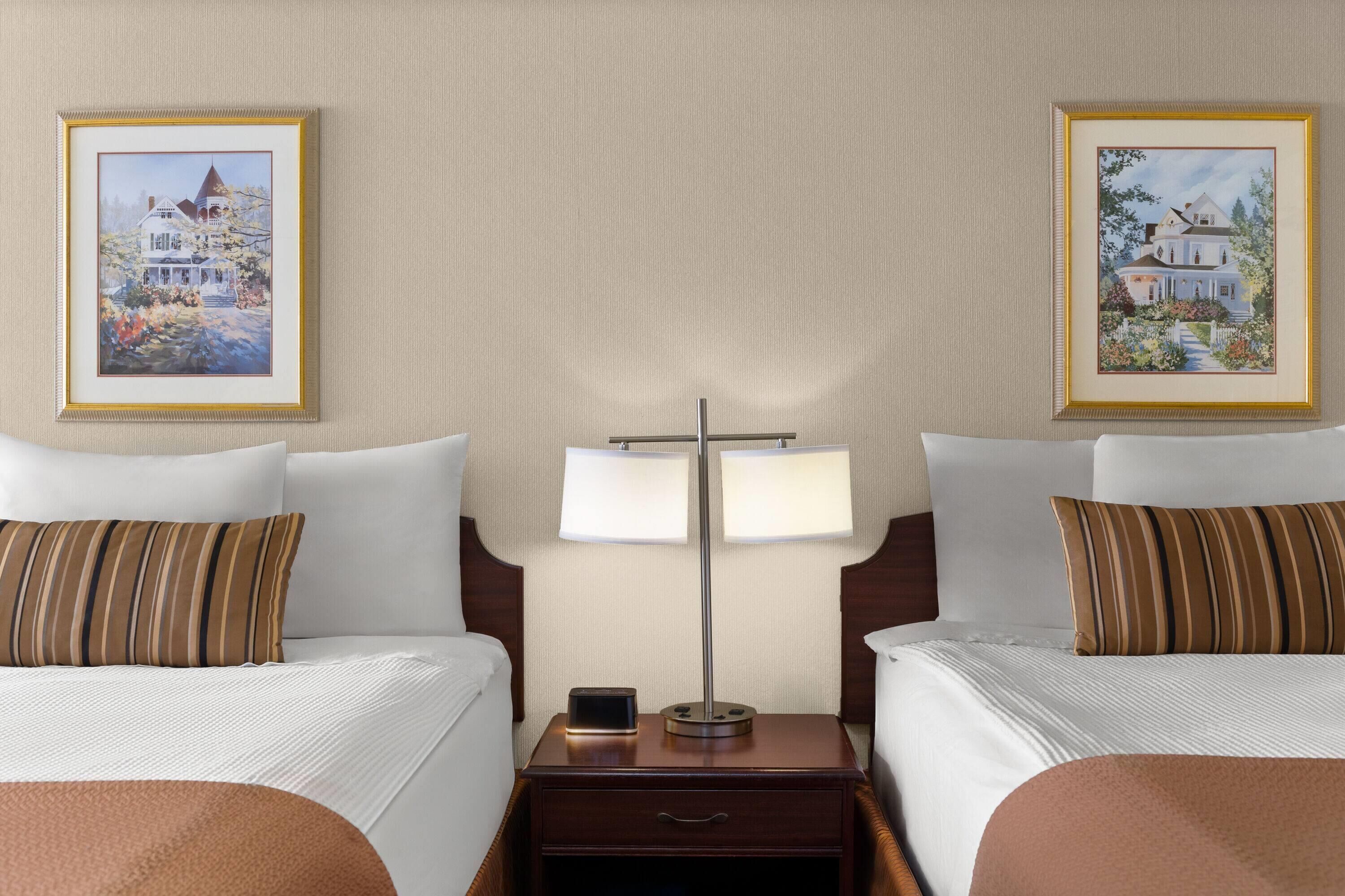 A hotel room with two beds, a nightstand with a lamp, and two framed paintings on the wall.
