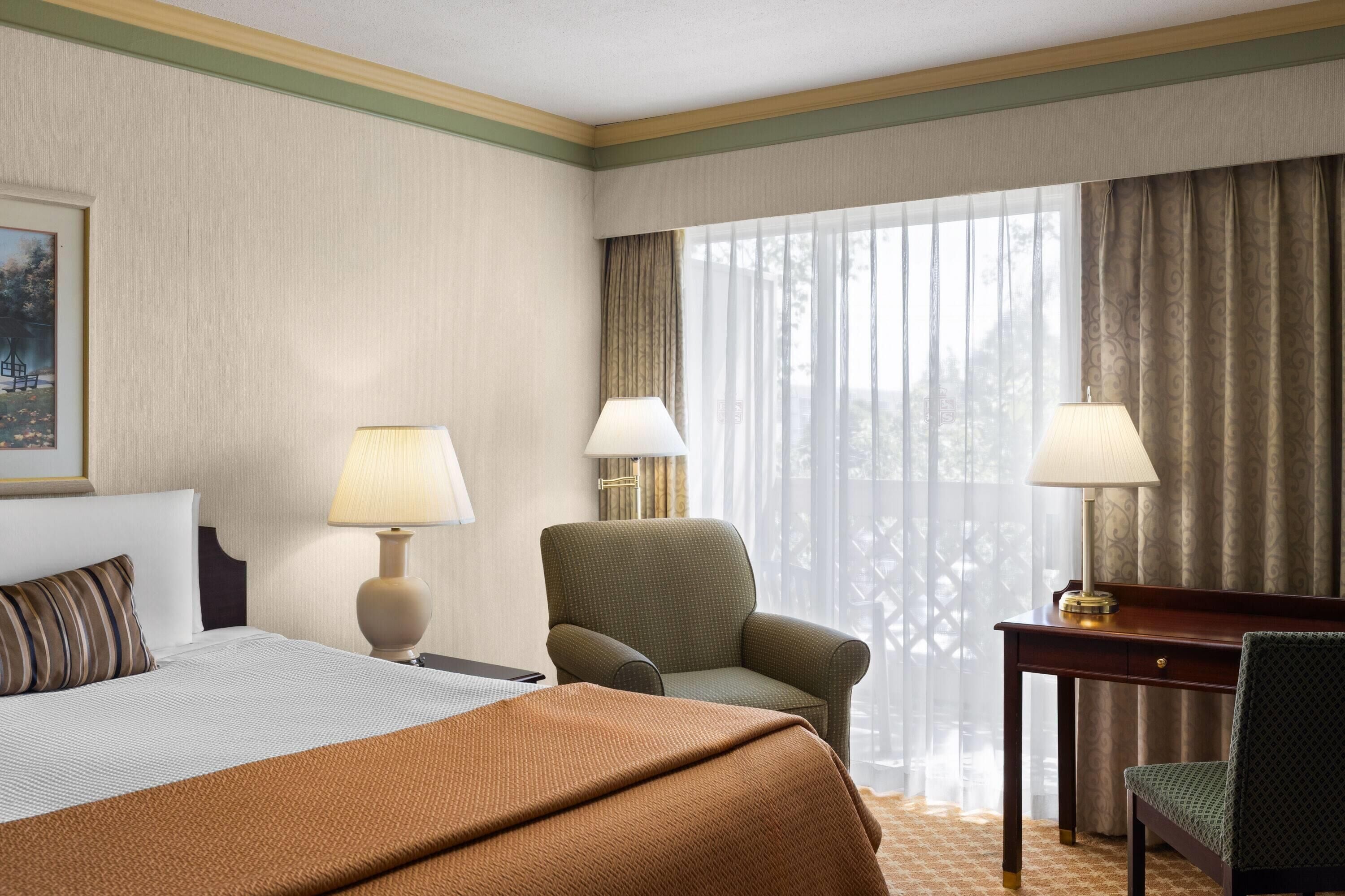 A hotel room with a bed, two chairs, a desk, lamps, and a large window with curtains.