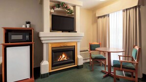 A hotel room with a fireplace, a television mounted above it, a small refrigerator, a microwave, a table with two chairs, and a large window with curtains. - Baymont by Wyndham Hinton (Hinton)