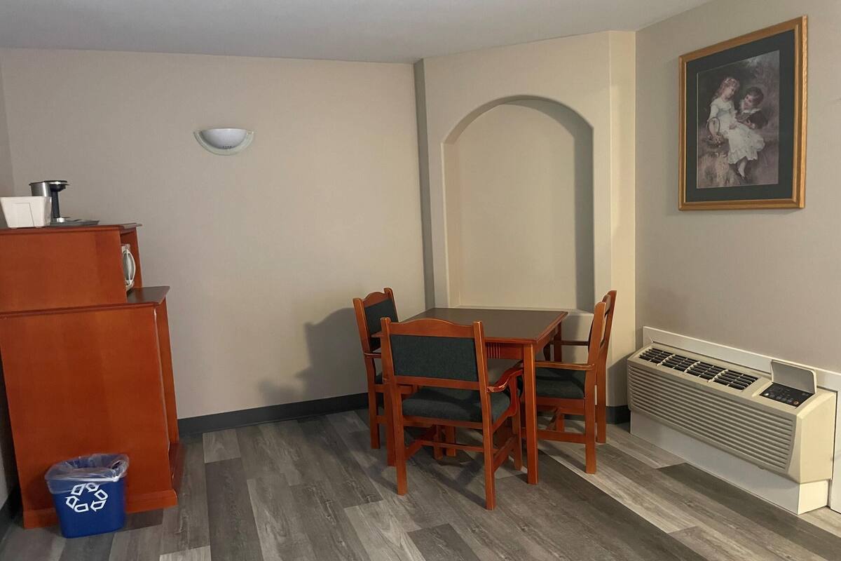 Un coin repas avec une table et des chaises, un petit réfrigérateur, une poubelle, une lampe fixée au mur, un radiateur et un tableau encadré accroché au mur.