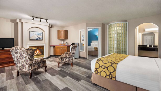 A hotel room with a fireplace, a bed with a patterned bedspread, a chair, and a desk with a television. - Baymont by Wyndham Hinton (Hinton)