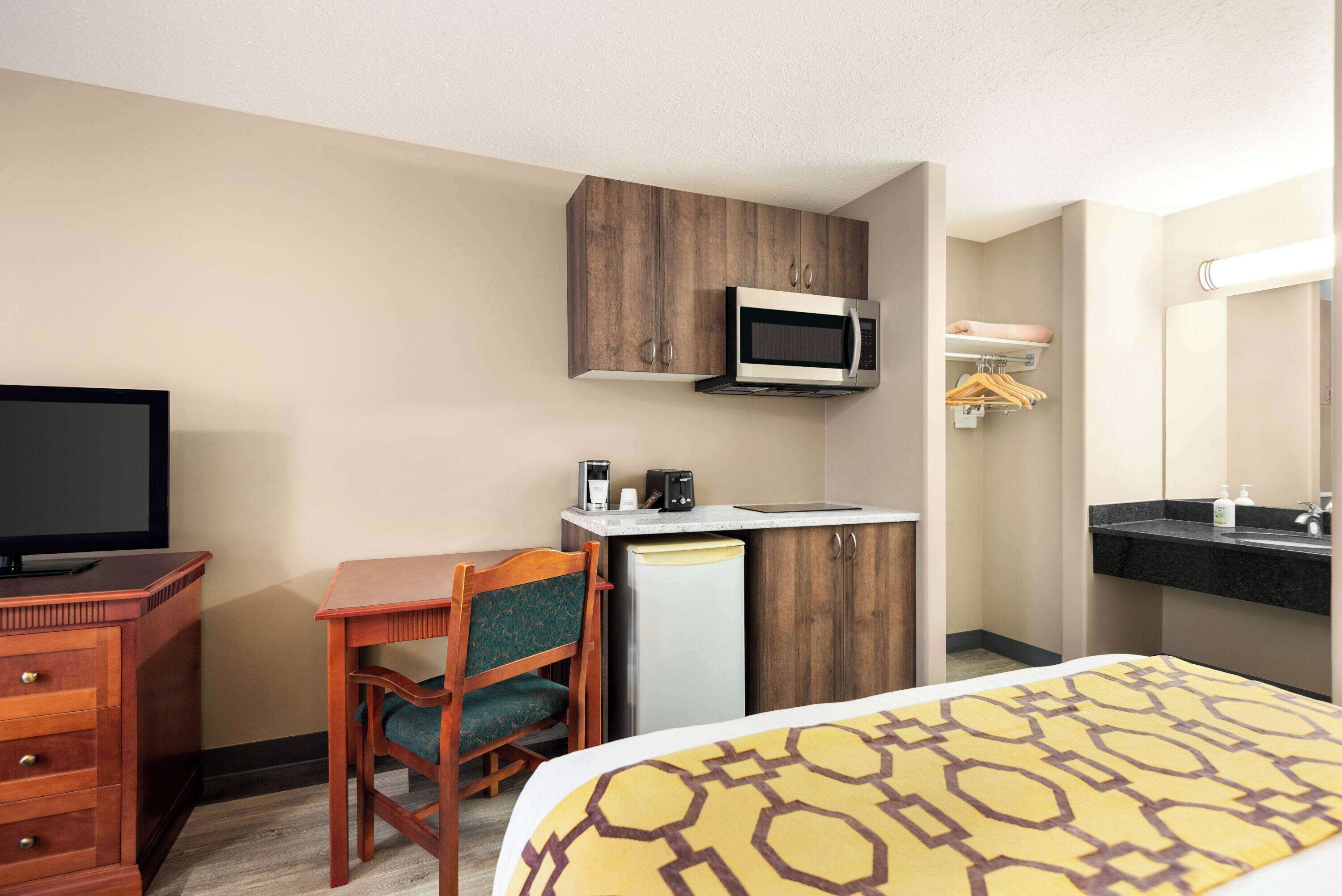 A hotel room with a bed, a desk with a chair, a microwave, a coffee maker, a refrigerator, a TV, and a bathroom with a sink and mirror.