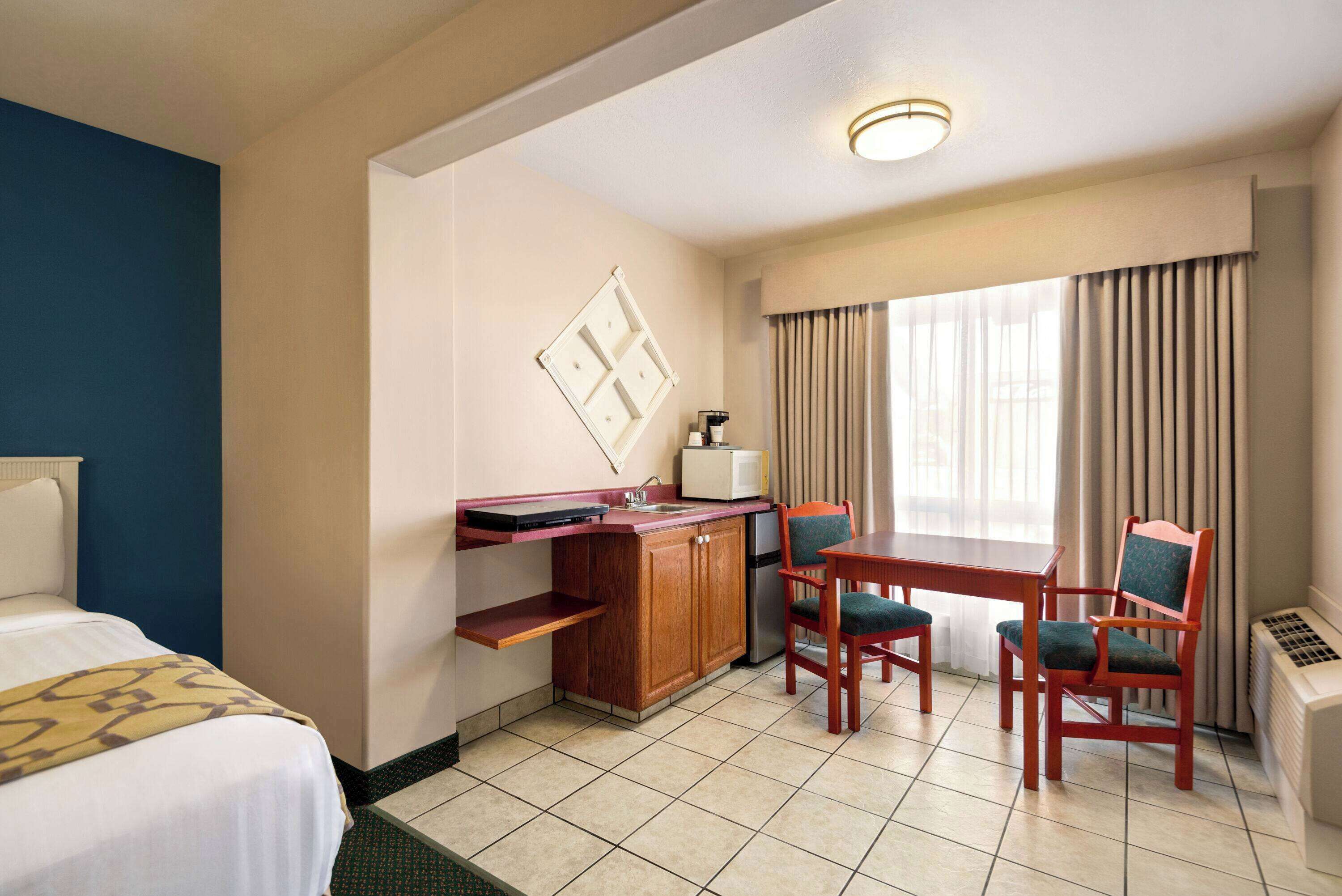A hotel room with a bed, a kitchenette, a dining area with two chairs, a microwave, and a window with curtains.