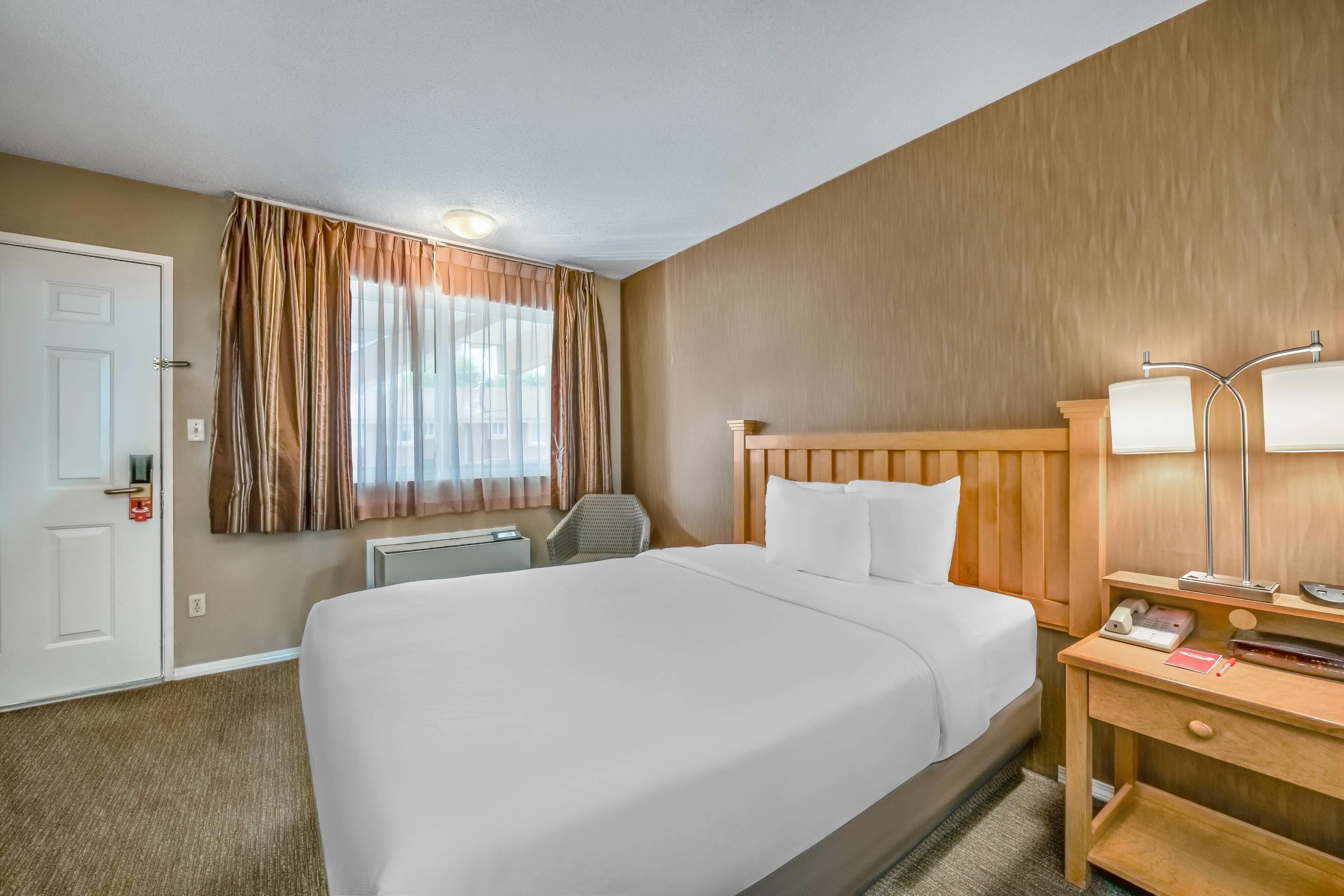 Ein Hotelzimmer mit einem großen Bett, einem hölzernen Kopfteil, einem Schreibtisch mit Lampe, einem Sessel und einem Fenster mit Vorhängen.