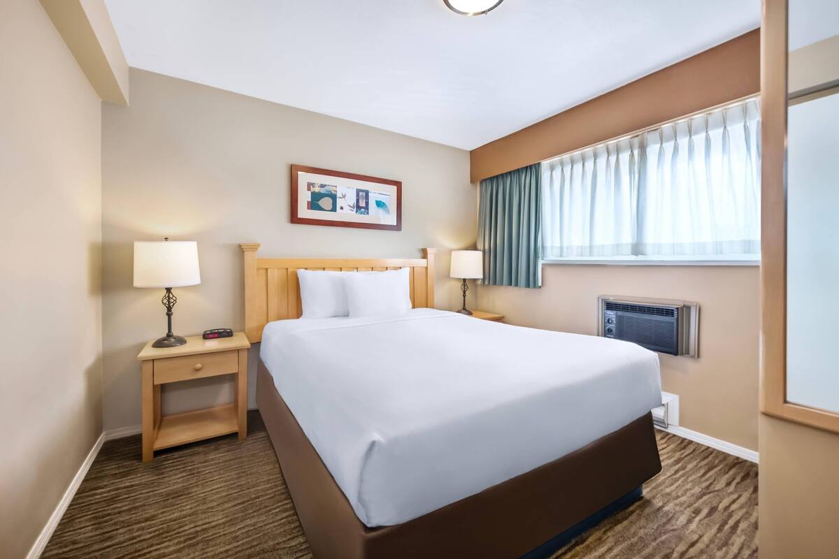 A hotel room with a large bed, two bedside tables with lamps, a wall-mounted air conditioner, and a window with curtains.
