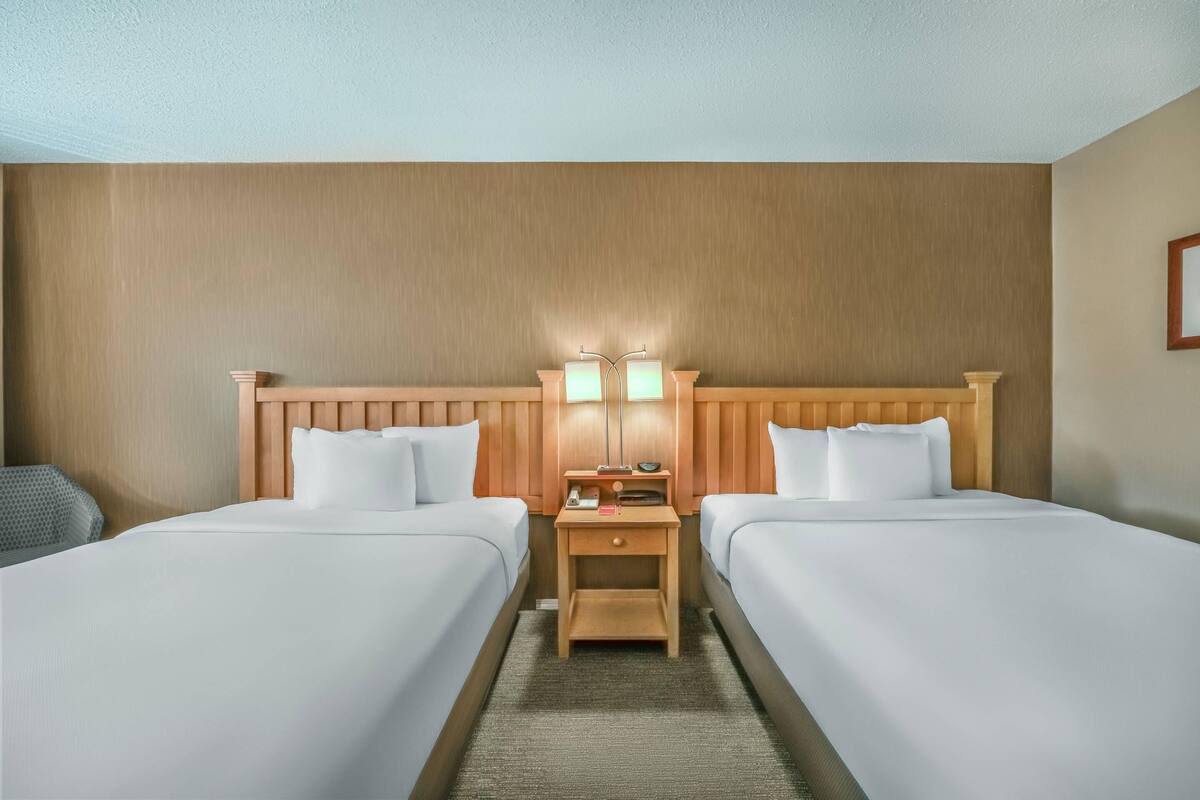 A hotel room with two beds, a wooden headboard, a nightstand with lamps, and a small table.