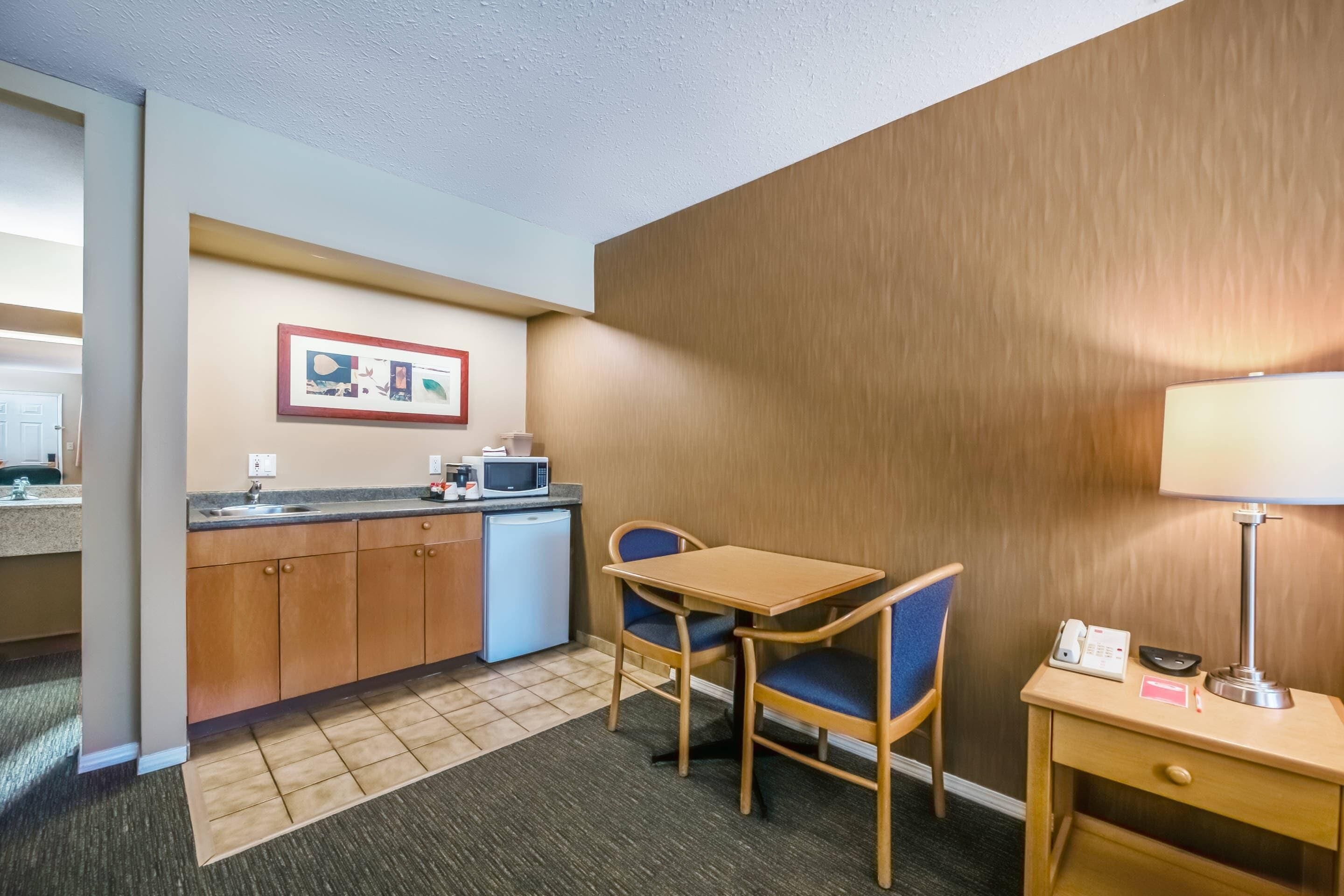 A compact hotel room with a kitchenette, a small dining table with two chairs, a bedside lamp, and a telephone.
