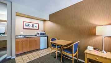 A compact hotel room with a kitchenette, a small dining table with two chairs, a bedside lamp, and a telephone.