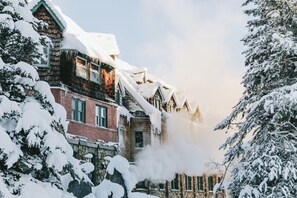 A snow-covered building with a chimney emitting smoke, surrounded by snow-laden trees. - Deer Lodge (Lake Louise)