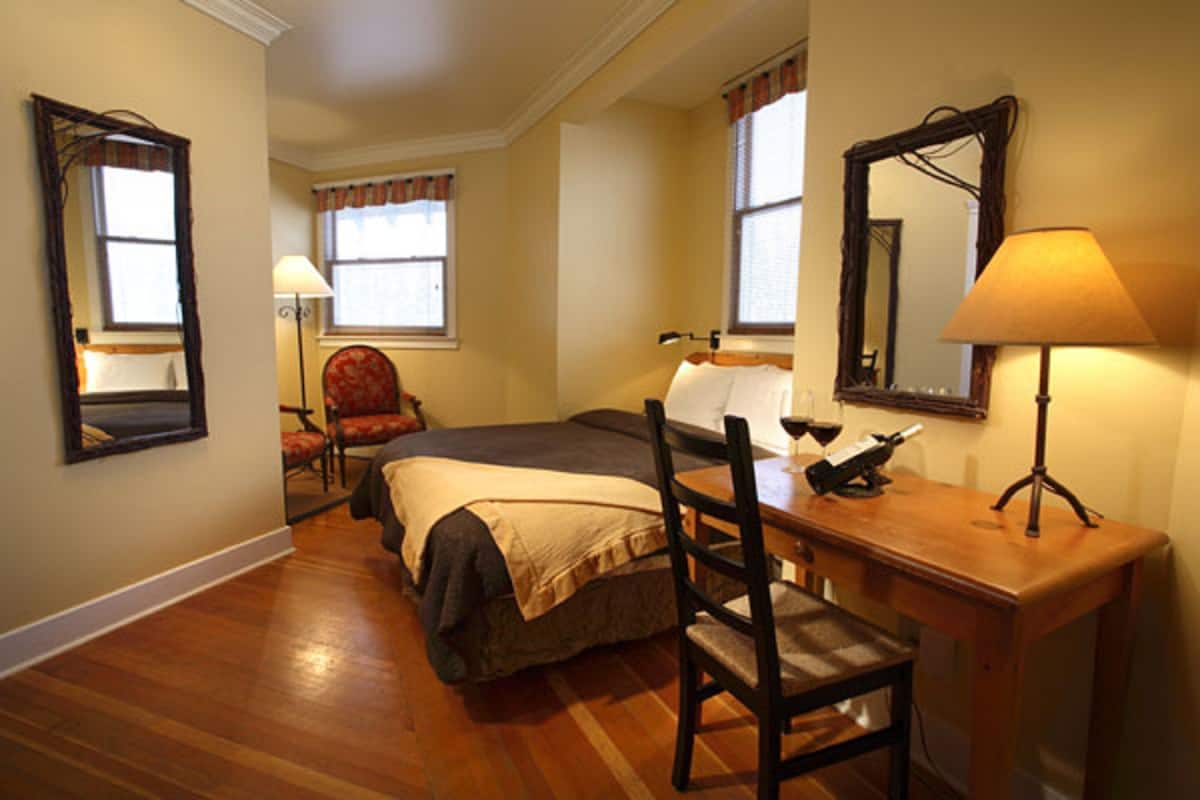 A hotel room with a bed, a desk with a lamp, a chair, and two windows with curtains.
