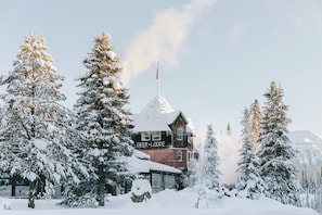 A snow-covered lodge with a prominent chimney, surrounded by evergreen trees. - Deer Lodge (Lake Louise)