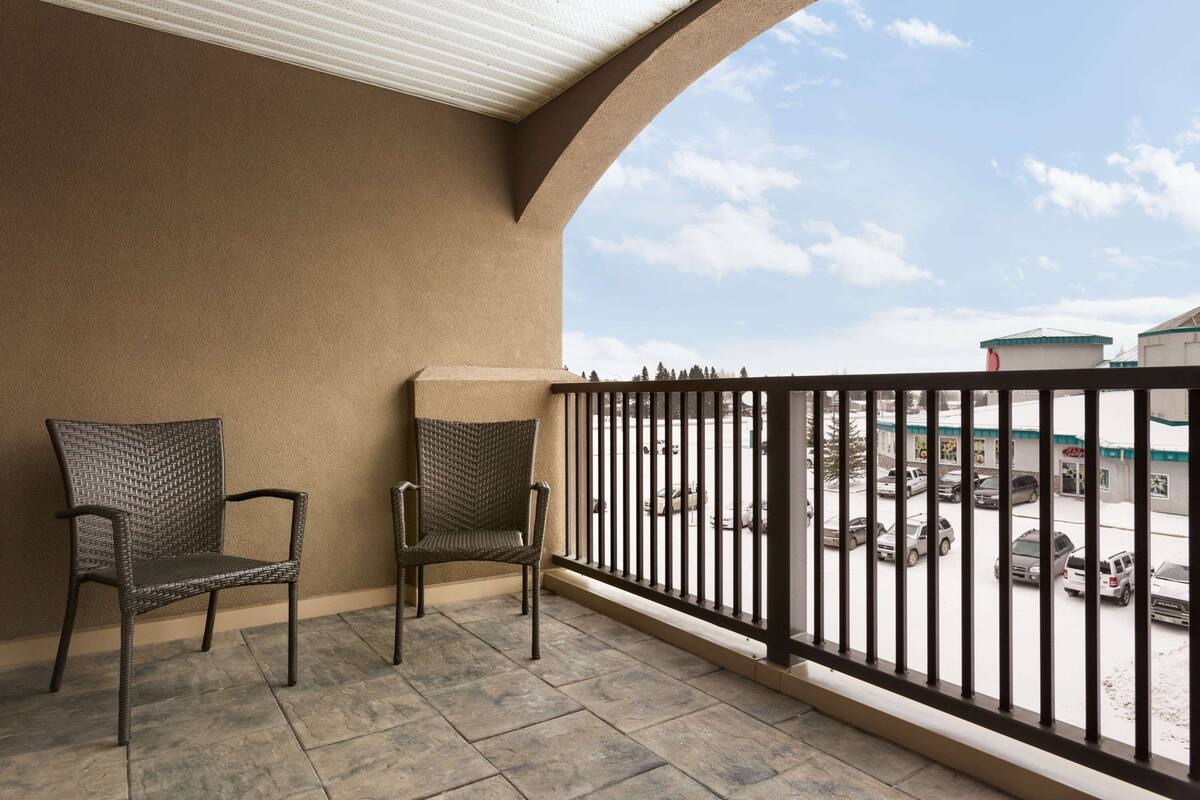 Un balcon avec deux fauteuils en osier, une rambarde en métal et une vue sur un parking et des bâtiments en arrière-plan.