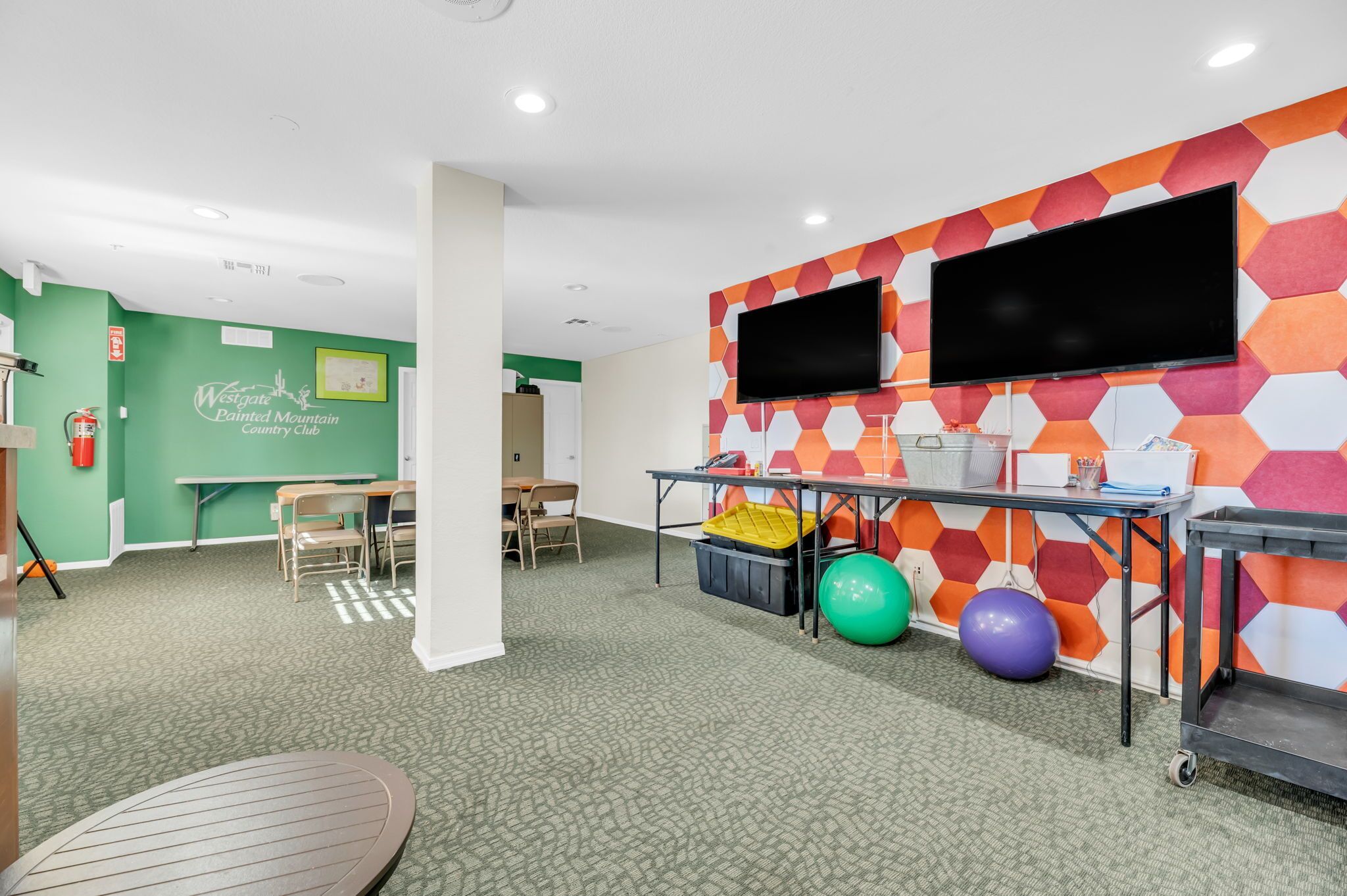 A room with a green wall, a table with chairs, a television mounted on a colorful hexagonal wall, and exercise balls.