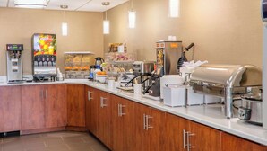 A self-service breakfast area with a coffee machine, juice dispenser, and various food items.
