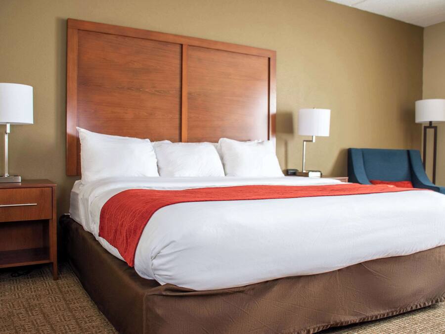 A hotel room with a large bed, two bedside tables, a nightstand, a red throw blanket, and two lamps.