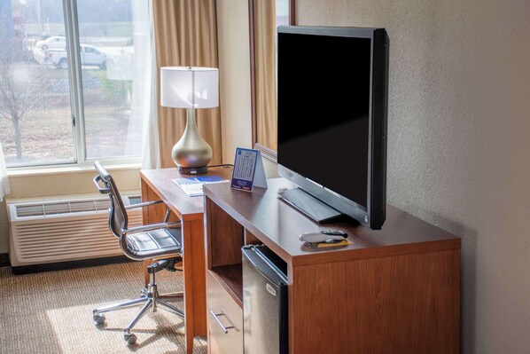 Habitación de hotel con un escritorio de madera, un televisor de pantalla plana, una lámpara, una silla y vistas a coches estacionados afuera.
