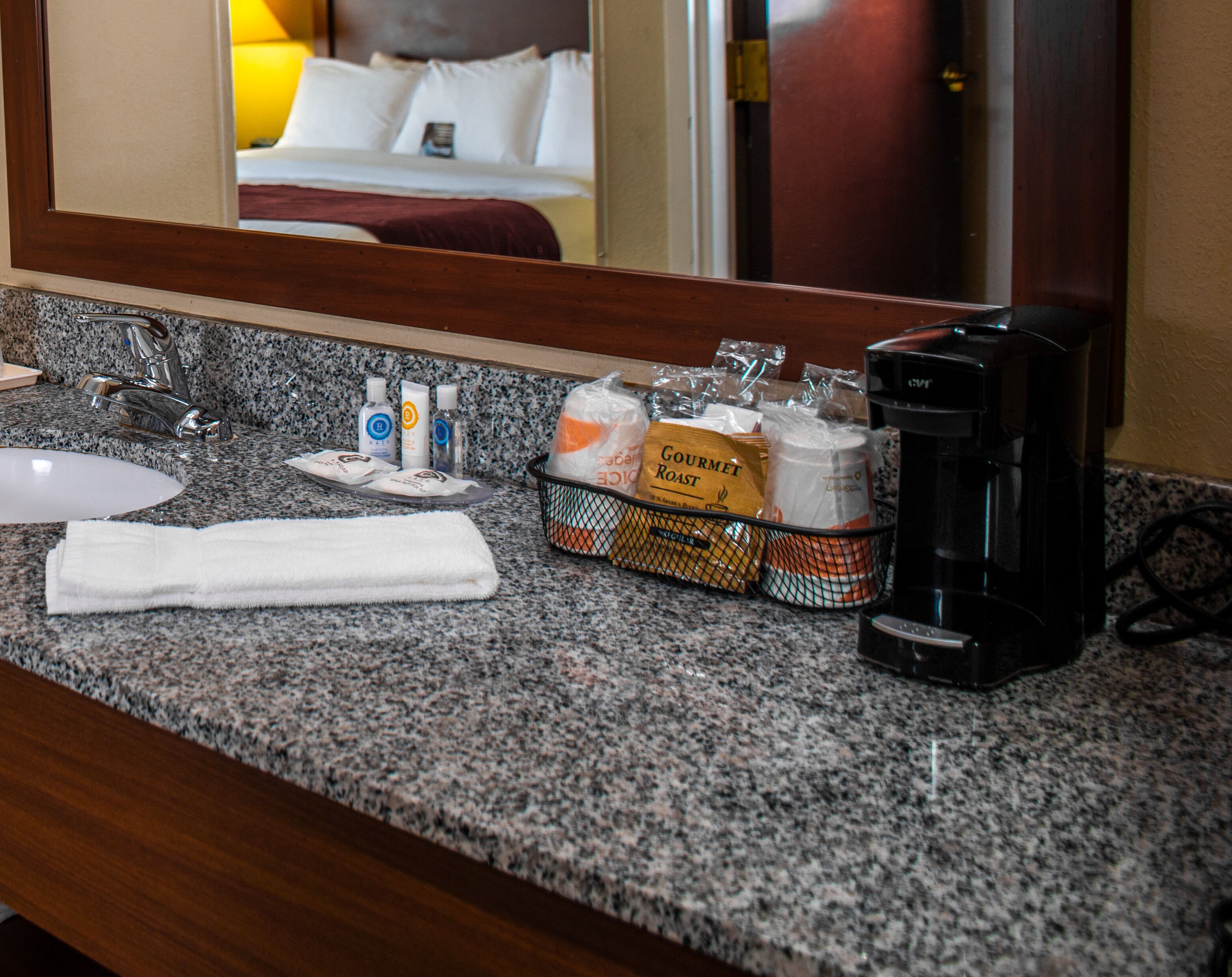 Baño de hotel con encimera de granito, cafetera, artículos de aseo y una cesta de galletas Gourmet Roast.