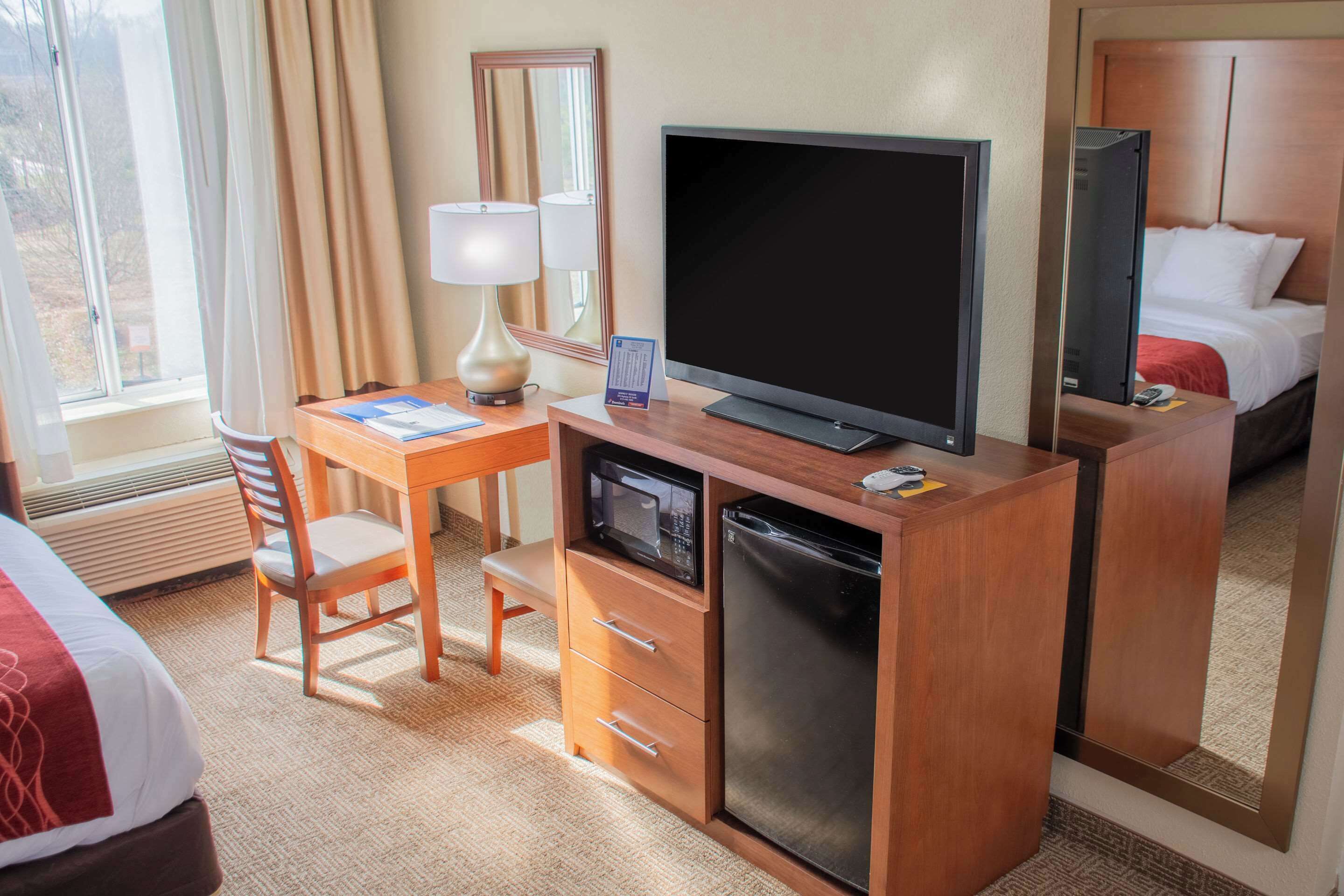 Habitació d'hotel amb escriptori de fusta, televisió, microones, nevera, llit i finestra amb cortines.