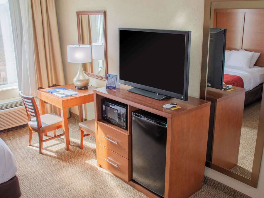 Habitació d'hotel amb escriptori de fusta, televisió, microones, nevera, llit i finestra amb cortines.