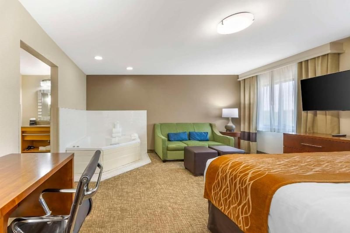 A hotel room with a bed, a green sofa, a desk, a chair, a bathtub, and a television.
