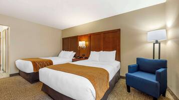 A hotel room with two beds, a blue armchair, and a nightstand with a lamp.