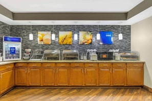 A modern kitchen with wooden cabinets, a granite countertop, and a backsplash with images of food. There are various kitchen appliances and a buffet setup.