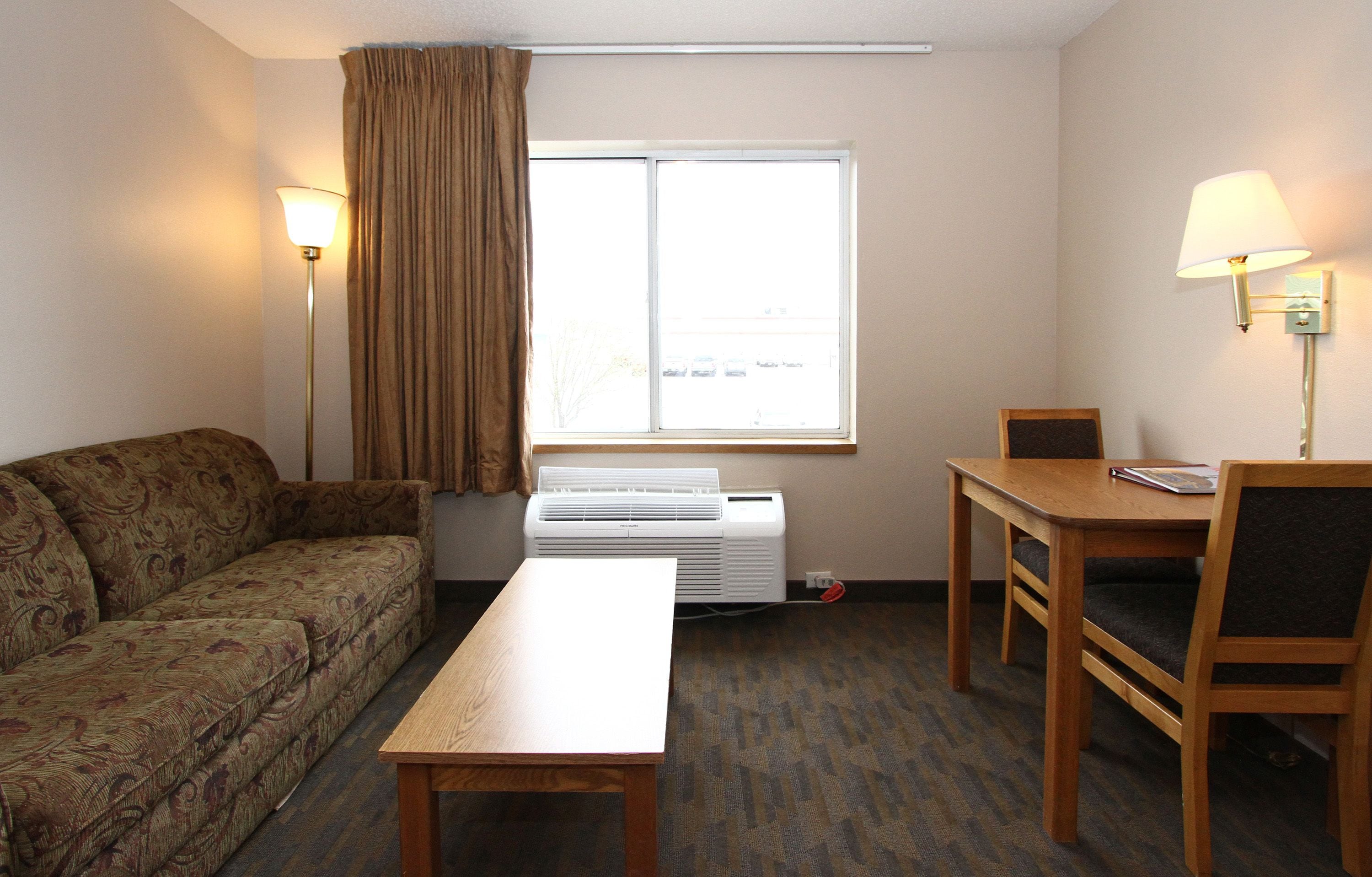 A hotel room with a sofa, a table, a chair, a window with curtains, and two wall lamps.