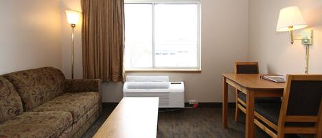 A hotel room with a sofa, a table, a chair, a window with curtains, and two wall lamps.