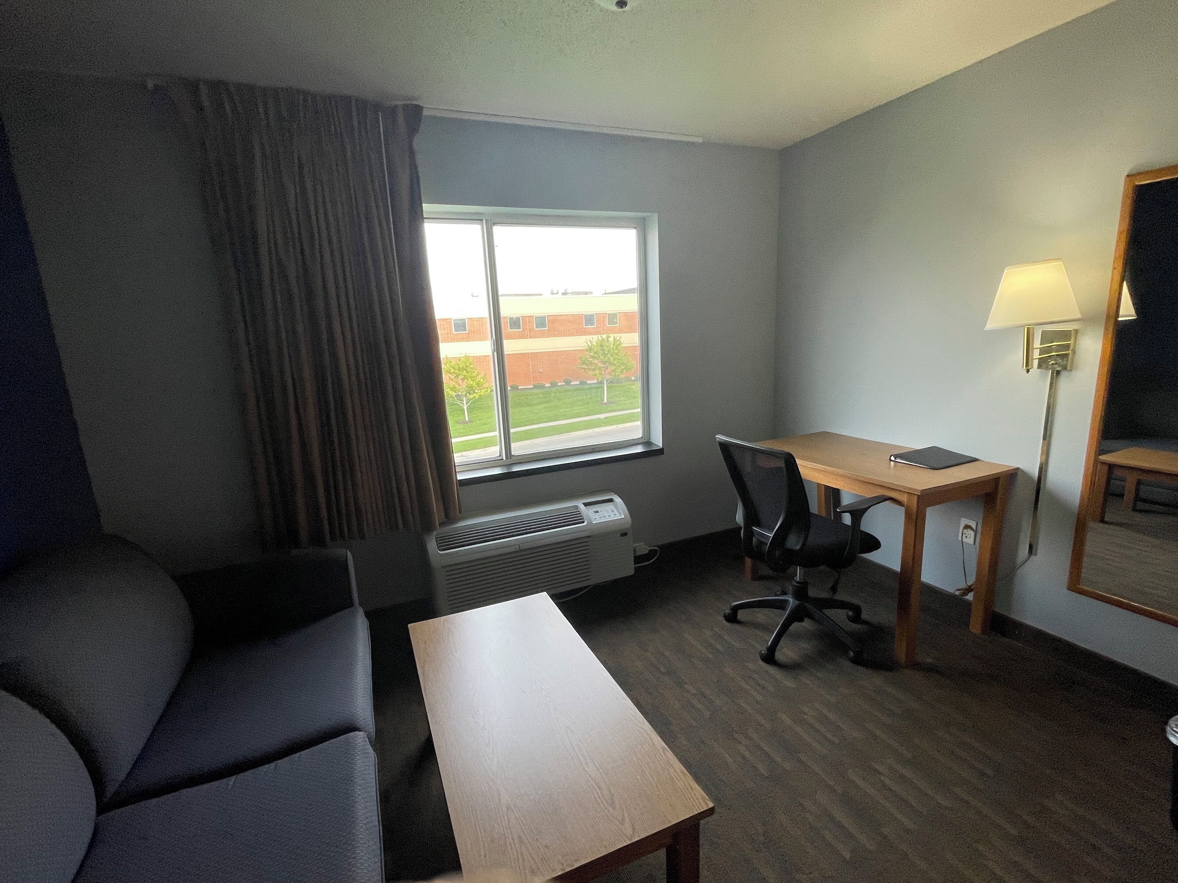 Habitación de hotel con cama, escritorio, silla, lámpara y ventana con vista a un edificio y árboles.