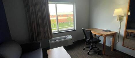 Habitación de hotel con cama, escritorio, silla, lámpara y ventana con vista a un edificio y árboles.